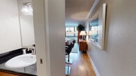 This single-floor studio is a well-appointed, efficiently designed space that makes excellent use of its layout. The main living and sleeping area is anchored by a statement bed with a tufted charcoal headboard set against a deep navy accent wall, creating a bold focal point. Rich warm-toned wood built-in cabinetry lines one wall, incorporating a built-in desk workspace with a black countertop — ideal for productivity. Hardwood flooring flows throughout, adding warmth and continuity across the space. Large windows wrap around the room, inviting abundant natural light and offering views of the surrounding neighborhood. A sleek media console with a flat-screen TV and a tall indoor plant complete the living area, contributing to a relaxed yet curated ambiance. The compact galley kitchen is fitted with stainless steel appliances, including a range, microwave, and refrigerator, all complemented by warm wood cabinetry. The bathroom features a wood vanity with a dark stone countertop, an undermount sink with brushed nickel fixtures, a shower-tub combination, and clean white finishes throughout. Crown molding and a textured ceiling add architectural character to the overall space.