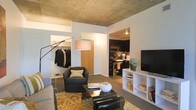 This single-floor space showcases a thoughtfully designed open-concept layout that blends industrial and modern aesthetics. The most striking architectural feature is the exposed concrete ceiling, which lends a raw, urban character to the interior while adding a sense of depth and texture overhead.

The living area is comfortably arranged with a cushioned armchair accented by a decorative pillow, a low-profile coffee table, and a plush area rug with an earthy, patterned design. A sleek white open shelving unit serves as an entertainment center, housing a flat-screen television and decorative objects. A large arched floor lamp provides warm ambient lighting to the space.

Visible through an open doorway, the kitchen features dark cabinetry, stainless steel appliances including a range and microwave, and a compact yet functional layout suited for everyday cooking. The dark finishes of the kitchen contrast effectively with the lighter tones of the living area.

An open closet area is also accessible from the main living space, offering organized hanging storage for clothing. The overall ambiance is modern and urban, with a neutral color palette complemented by warm accent pieces throughout.