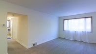 This single-floor space presents a straightforward and functional layout centered around a spacious main living area. The room features wall-to-wall carpeting in a neutral beige tone, providing a soft underfoot feel that complements the clean, white walls throughout. A wood-trimmed window allows natural light to filter in through sheer white curtains, brightening the interior and lending an open, airy quality to the space. The walls are smooth and painted in a crisp white, creating a blank canvas well-suited for a variety of interior styles. A recessed opening on one wall suggests a passageway or adjoining area, adding to the functional flow of the layout. Baseboard trim runs cleanly along the perimeter of the room, and a floor-level heating vent is positioned along the base of the wall, indicating a built-in climate control system. The overall ambiance is calm and neutral, offering a versatile foundation for furnishing and décor. The space is well-proportioned, providing ample room for living and everyday use, and its simple, uncluttered design makes it easy to envision a range of arrangements and uses.
