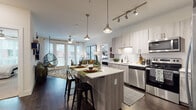 This well-appointed single-floor residence offers a thoughtfully designed open-concept layout that seamlessly connects the kitchen, dining, and living areas. The kitchen is a standout feature, showcasing light wood-grain cabinetry, white quartz countertops, a full suite of stainless steel appliances, a subway tile backsplash, and a central island with bar seating — ideal for both casual dining and entertaining. Track lighting and pendant fixtures provide layered illumination throughout the space. Dark hardwood flooring runs through the main living areas, adding warmth and contrast to the light cabinetry and crisp white walls. The living room is bright and inviting, benefiting from generous natural light through full-height glass doors that open to a private balcony, complemented by a ceiling fan overhead. The two bedrooms are carpeted for comfort, each offering ample space and recessed lighting. The primary bathroom features a dual-sink vanity with wood-toned cabinetry and quartz surfaces, alongside a glass-enclosed shower. The secondary bathroom includes a matching vanity and a shower-tub combination. Neutral tones and clean lines define the overall aesthetic, creating a cohesive, contemporary ambiance throughout the residence.