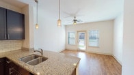 This single-floor unit offers a thoughtfully designed layout with distinct living, kitchen, bedroom, and bathroom spaces. The kitchen serves as a central highlight, featuring dark espresso cabinetry, a subway tile backsplash, stainless steel appliances including a refrigerator, range, and microwave, and a curved granite-topped peninsula with a double undermount sink. Decorative pendant lighting above the peninsula adds character, while track lighting illuminates the workspace efficiently. The kitchen opens directly to the entryway and living area, both finished with warm wood-style hard flooring that enhances the sense of continuity. The living area is bright and airy, benefiting from a ceiling fan, a glass exterior door, and flanking windows that invite natural light and outdoor views. The bedroom offers plush carpeting, a ceiling fan with light, and a large window with exterior views. The bathroom is well-equipped with a granite-topped vanity featuring rich wood cabinetry, a framed mirror, and a full tub-shower combination with tile surround and chrome fixtures. Light tile flooring completes the bathroom's clean, functional aesthetic.
