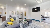 This contemporary single-floor amenity space offers a thoughtfully designed layout that combines social, functional, and fitness-oriented areas into one cohesive environment. The main lounge serves as the centerpiece, featuring gleaming marble-look tile flooring, bold blue baseboards, and striking circular ring chandeliers that lend a modern, upscale ambiance. Plush gray sectional sofas, glass-top coffee tables, and yellow accent pillows create an inviting atmosphere, while a wall-mounted flat-screen TV and open shelving units add both function and style. Curated wall art and decorative elements throughout elevate the aesthetic. French doors with views of greenery flood the lounge with natural light and provide a seamless connection to the outdoors. A dedicated meeting room offers a more intimate setting, appointed with warm hardwood-style flooring, a striking round marble-top table, and sleek white chairs — ideal for small gatherings or focused work sessions. The fitness center is well-appointed with rubber flooring, full-wall mirrors, and a comprehensive range of equipment including a treadmill, stationary bike, power rack, free weights, and exercise mats. Together, these spaces form a versatile and polished amenity hub designed to support both relaxation and an active lifestyle.