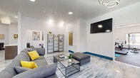 This contemporary single-floor amenity space offers a thoughtfully designed layout that combines social, functional, and fitness-oriented areas into one cohesive environment. The main lounge serves as the centerpiece, featuring gleaming marble-look tile flooring, bold blue baseboards, and striking circular ring chandeliers that lend a modern, upscale ambiance. Plush gray sectional sofas, glass-top coffee tables, and yellow accent pillows create an inviting atmosphere, while a wall-mounted flat-screen TV and open shelving units add both function and style. Curated wall art and decorative elements throughout elevate the aesthetic. French doors with views of greenery flood the lounge with natural light and provide a seamless connection to the outdoors. A dedicated meeting room offers a more intimate setting, appointed with warm hardwood-style flooring, a striking round marble-top table, and sleek white chairs — ideal for small gatherings or focused work sessions. The fitness center is well-appointed with rubber flooring, full-wall mirrors, and a comprehensive range of equipment including a treadmill, stationary bike, power rack, free weights, and exercise mats. Together, these spaces form a versatile and polished amenity hub designed to support both relaxation and an active lifestyle.