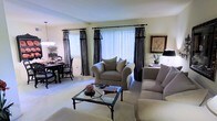 This single-floor residence offers a thoughtfully arranged living space with a warm and inviting ambiance. The living room serves as the centerpiece of the home, featuring plush, neutral-toned upholstered seating arranged around a glass-top coffee table with a wrought iron frame, lending the room a classic yet refined aesthetic. Large windows dressed with floor-length patterned drapery in dark tones allow natural light to fill the room, complemented by vertical blinds that offer adjustable light control. The soft carpeted flooring runs throughout, adding warmth and continuity to the space. Adjacent to the living area, a formal dining space accommodates a dark wood dining table and matching chairs, ideal for everyday meals or hosting guests. Decorative accents, including framed artwork and potted greenery, contribute to the polished, lived-in character of the home. The overall color palette is neutral and calming, with light walls providing a versatile backdrop for the furnishings. The open flow between the living and dining areas enhances the sense of space, making this residence feel both functional and comfortable for everyday living.
