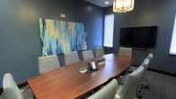 This professional conference room offers a refined and focused environment ideal for meetings and collaborative sessions. The space is anchored by a substantial oval-shaped wood conference table with a rich, warm finish, surrounded by a set of matching gray upholstered chairs that provide both comfort and a cohesive aesthetic. Deep blue-gray walls lend the room a calm, authoritative atmosphere, complemented by a striking large-scale abstract artwork in coordinating shades of blue and cream that serves as a bold focal point. A flat-screen display mounted on the wall provides audiovisual capabilities for presentations, while a conference phone sits centrally on the table for seamless communication. Natural light filters in through a window fitted with blinds, adding brightness without distraction. An elegant geometric chandelier overhead enhances the room's refined character. Recessed lighting supplements the ambient glow, ensuring the space is well-lit throughout. The single-floor layout is efficiently designed, making the most of the available footprint to create an intimate yet fully functional boardroom setting suitable for a range of professional engagements.
