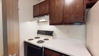 The kitchen showcases a classic, functional design anchored by rich dark wood cabinetry that extends to the upper walls, providing ample storage space. The upper cabinets feature a warm, wood-grain finish that contrasts neatly with the crisp white walls, creating a traditional aesthetic. A built-in range hood is mounted beneath the upper cabinets, positioned directly above the freestanding electric range. The range features four coil burners set into a white cooktop surface, paired with a black body and digital controls, offering a straightforward cooking setup. Adjacent to the range, light-toned speckled countertops provide a clean workspace, complementing the overall color palette of the kitchen. The lower cabinetry matches the upper units, maintaining a cohesive look throughout the space. The white walls reflect light effectively, contributing to a bright and practical cooking environment. The layout is compact yet thoughtfully arranged, ensuring that cooking essentials are within easy reach. Overall, the kitchen balances utility with a timeless, no-frills design approach.