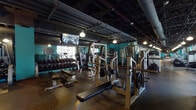 This fitness center is a comprehensively appointed gym spread across a single expansive floor, designed to accommodate a full range of workout disciplines. The space is defined by its industrial-chic aesthetic, with exposed black ceilings revealing ductwork, pipes, and pendant lighting that casts a warm, focused glow throughout.

The strength training area is generously outfitted with multiple power racks, barbells, weight plates, benches, and a full rack of dumbbells, all set against floor-to-ceiling mirrors that enhance the sense of depth and allow users to monitor their form. Teal-painted accent walls provide bold, energizing contrast against the dark ceiling and speckled rubber flooring.

The cardio zone features a long row of treadmills and elliptical machines positioned alongside large windows that frame urban cityscape views, filling the area with natural light. Stationary bikes and additional cardio equipment line the opposite wall, which is adorned with a vibrant, large-scale motivational mural — a striking visual anchor for the space.

Adjacent to the cardio area, a dedicated hardwood-floored studio zone offers open space ideal for stretching, functional training, or group exercise. Together, the zones flow seamlessly, creating a dynamic and motivating environment well-suited for diverse fitness routines.