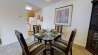 Located at 310 N Broad St in Carneys Point, New Jersey, this single-floor residence offers a thoughtfully arranged living environment designed for everyday comfort. The dining area serves as a central gathering space, accommodating a full-sized dining table with upholstered seating, and is accented by decorative wall art that adds a warm, homey character to the room. Soft carpeting underfoot contributes to the cozy atmosphere throughout the living spaces. Adjacent to the dining area, the kitchen is efficiently laid out with ample cabinetry finished in warm wood tones, providing generous storage. White appliances, including a full-sized refrigerator, complement the clean and functional design of the kitchen. The transition between the dining and kitchen areas flows naturally, making the space feel connected and easy to navigate. The neutral wall palette throughout the residence creates a bright and open feel, allowing the décor and furnishings to stand out. The overall layout reflects a practical single-floor design that prioritizes ease of movement and livability, making it well-suited for a variety of lifestyles.
