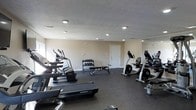 This fitness center offers a practical and well-organized space designed to accommodate a variety of workout needs. The room features a dark, durable rubberized floor that provides both safety and comfort during exercise. Recessed lighting is evenly distributed across the textured ceiling, ensuring the space is well-lit throughout. The neutral-toned walls contribute to a clean, focused atmosphere conducive to exercise.

The cardio section is prominently featured along one side of the room and includes multiple treadmills and an elliptical machine, providing a range of options for cardiovascular training. On the opposite side, upright and recumbent exercise bikes offer additional low-impact cardio alternatives. A dedicated free weights area is centrally positioned, featuring a multi-tiered dumbbell rack stocked with a variety of weights, accompanied by a flat weight bench for strength and resistance training exercises. A large mirror along one wall enhances the sense of space while allowing users to monitor their form. A single access door provides entry to the room, keeping the layout efficient and uncluttered. Overall, this fitness center is thoughtfully arranged to maximize functionality within its footprint.