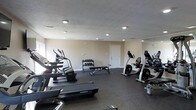 This fitness center offers a practical and well-organized space designed to accommodate a variety of workout needs. The room features a dark, durable rubberized floor that provides both safety and comfort during exercise. Recessed lighting is evenly distributed across the textured ceiling, ensuring the space is well-lit throughout. The neutral-toned walls contribute to a clean, focused atmosphere conducive to exercise.

The cardio section is prominently featured along one side of the room and includes multiple treadmills and an elliptical machine, providing a range of options for cardiovascular training. On the opposite side, upright and recumbent exercise bikes offer additional low-impact cardio alternatives. A dedicated free weights area is centrally positioned, featuring a multi-tiered dumbbell rack stocked with a variety of weights, accompanied by a flat weight bench for strength and resistance training exercises. A large mirror along one wall enhances the sense of space while allowing users to monitor their form. A single access door provides entry to the room, keeping the layout efficient and uncluttered. Overall, this fitness center is thoughtfully arranged to maximize functionality within its footprint.