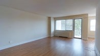This single-floor residence offers a clean, open layout designed for comfortable everyday living. The main living area is immediately welcoming, characterized by warm hardwood flooring that extends throughout the space, creating a sense of continuity and warmth underfoot. Neutral, light-toned walls provide a versatile backdrop that complements a wide range of furnishing styles.

Natural light is a defining feature of the living area, which benefits from a large dual-panel window paired with a sliding glass door that opens directly onto a private balcony. This combination allows for an abundance of daylight to fill the room while offering views of the surrounding greenery visible through the glass. The balcony access also provides a seamless indoor-outdoor connection, ideal for enjoying fresh air.

A baseboard heating unit beneath the window ensures the space remains comfortable year-round. The overall design is understated and functional, with smooth ceilings and clean white baseboards that lend the interior a well-maintained, polished appearance. The open feel of the living area makes it highly adaptable, offering ample room for a variety of furniture arrangements to suit different lifestyle needs. The space presents a bright, airy ambiance throughout.