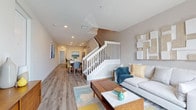 This two-story home offers a thoughtfully designed layout that balances open communal spaces with private retreats across two levels. The main floor centers around an open-concept living, dining, and kitchen area unified by warm wood-look flooring and recessed lighting throughout. The living room provides a welcoming gathering space anchored by a comfortable sofa, colorful area rug, and a crisp white staircase with classic spindle railings that serves as an architectural focal point. The kitchen is well-appointed with white shaker cabinetry, stainless steel appliances including a refrigerator with ice maker, a range with microwave, and a tasteful tile backsplash. A peninsula with bar seating flows seamlessly into the dining area, making the space ideal for both everyday meals and entertaining. A full bathroom on the main level features clean white finishes, a single vanity, and a tub-shower combination with wood-look flooring. Upstairs, the second floor hosts the bedrooms, accessible via the central staircase. The primary bedroom, visible in the dollhouse view, is generously proportioned and bright. Throughout the home, neutral tones, modern hardware, and consistent flooring choices create a cohesive, contemporary aesthetic.