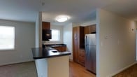 Located at 26 Cheswold Boulevard in Newark, Delaware, this single-floor residence offers a thoughtfully designed layout with a clean, modern aesthetic. The kitchen serves as a central highlight of the space, showcasing rich, warm-toned wood cabinetry that extends from floor to ceiling, providing generous storage throughout. Dark granite-style countertops offer a striking contrast against the lighter walls, while a breakfast bar peninsula creates a natural divide between the kitchen and the adjacent living area — ideal for casual dining or entertaining. Stainless steel appliances, including a full-size refrigerator, complement the contemporary finish of the space. A built-in range and a stainless sink with a window above add both functionality and a sense of openness. The flooring transitions from warm hardwood in the kitchen and dining areas to carpet in the living spaces, adding comfort and visual variety. Recessed and flush-mount ceiling lighting ensures the space is well-lit throughout. The overall ambiance is modern and well-appointed, with a cohesive design language that balances practicality with style, making it a comfortable and functional living environment.