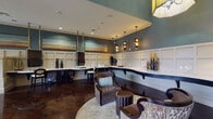 This sophisticated single-floor community clubhouse or leasing lounge offers a thoughtfully designed interior that balances functionality with upscale aesthetics. The space is anchored by rich, dark hardwood flooring that flows throughout, complemented by soaring ceilings that lend an open, airy feel to the entire area.

The main lounge area features elegant upholstered armchairs and side tables arranged atop a circular area rug, creating an inviting conversational setting. A striking yellow lantern-style pendant light serves as a standout focal point, adding warmth and character. The walls are adorned with white wainscoting panels and painted in a sophisticated teal-blue hue above, giving the space a refined yet welcoming atmosphere. Large-scale horizontal landscape paintings add artistic interest along the upper walls.

A dedicated business center lines one wall, offering a curved white countertop workstation with dark cabinetry below, multiple computer monitors suspended from the ceiling, and ample workspace seating. Nearby, a casual dining area provides a round dark wood table surrounded by green upholstered chairs, bathed in natural light from large windows with decorative Roman shades. A built-in banquette with striped upholstery offers additional seating. Glass partition doors connect the various zones, maintaining an open flow while defining distinct functional areas throughout the space.