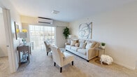 This single-floor residence at Gardens On Gateway Senior Apartments in McCordsville, Indiana, offers a thoughtfully designed layout suited for comfortable senior living. The living room serves as the heart of the home, featuring plush carpeting, a wall-mounted mini-split HVAC system, and a large sliding glass door that opens to a private balcony, filling the space with natural light and outdoor views. The open-concept kitchen is outfitted with warm-toned wooden cabinetry, granite-style countertops, stainless steel appliances including a refrigerator, range, microwave, and dishwasher, and a peninsula with bar seating. An adjacent dining area provides a welcoming spot for meals. Two well-sized bedrooms each feature carpet flooring, natural light from windows with blinds, and neutral wall tones, with one bedroom offering space for a desk or workspace. The bathroom is designed with accessibility in mind, featuring stone-look tile flooring, a wood-toned vanity, grab bars, and a walk-in shower with glass doors. Throughout, the residence maintains a calm, neutral palette that contributes to a serene and practical living environment.