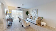 This single-floor residence at Gardens On Gateway Senior Apartments in McCordsville, Indiana, offers a thoughtfully designed layout suited for comfortable senior living. The living room serves as the heart of the home, featuring plush carpeting, a wall-mounted mini-split HVAC system, and a large sliding glass door that opens to a private balcony, filling the space with natural light and outdoor views. The open-concept kitchen is outfitted with warm-toned wooden cabinetry, granite-style countertops, stainless steel appliances including a refrigerator, range, microwave, and dishwasher, and a peninsula with bar seating. An adjacent dining area provides a welcoming spot for meals. Two well-sized bedrooms each feature carpet flooring, natural light from windows with blinds, and neutral wall tones, with one bedroom offering space for a desk or workspace. The bathroom is designed with accessibility in mind, featuring stone-look tile flooring, a wood-toned vanity, grab bars, and a walk-in shower with glass doors. Throughout, the residence maintains a calm, neutral palette that contributes to a serene and practical living environment.