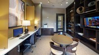 This single-floor business center is thoughtfully designed to support both focused work and collaborative meetings. The space is anchored by a dramatic floor-to-ceiling built-in shelving unit finished in dark espresso wood, housing a centrally mounted flat-screen television and an assortment of decorative sculptural objects, including metallic spheres and abstract art pieces, adding a refined, curated aesthetic.

Multiple computer workstations are arranged along a continuous countertop, each outfitted with a desktop computer and ergonomic task chair. Large windows along one wall allow natural light to filter into the workspace, complemented by warm pendant lighting and recessed ceiling fixtures that maintain a well-lit environment throughout.

A round dining-height table surrounded by contemporary molded chairs provides a flexible area for group discussions or informal meetings. A dedicated alcove houses a print and copy station with built-in dark cabinetry and a white countertop, keeping workflow efficient. Textured wall coverings in warm taupe and gold tones, along with large-scale framed artwork, contribute to a polished, professional ambiance throughout the space.
