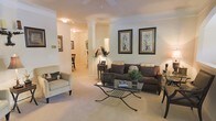 This single-floor residence exudes a classic and refined character throughout its thoughtfully designed layout. The living area serves as the focal point of the space, showcasing decorative columns that add an architectural elegance while subtly defining the room without closing it off. Soft, neutral-toned carpet flows underfoot, complementing the warm, cream-colored walls and contributing to the space's welcoming atmosphere. The living room is generously proportioned, comfortably accommodating a rich, dark brown sofa paired with coordinating accent chairs upholstered in a subtle pattern. A glass-topped coffee table with wrought iron legs anchors the seating arrangement, adding a touch of sophistication. A pair of framed botanical prints adorns the wall, lending a tasteful, curated aesthetic. Warm overhead lighting illuminates the space evenly, enhancing the cozy ambiance. Through the columns, a glimpse into an adjoining corridor and dining area reveals a connected, open flow between living spaces. A decorative accent table with an ornamental lamp and greenery arrangement adds a welcoming touch near the entryway. Crown molding along the ceiling further elevates the interior's polished, traditional character throughout.