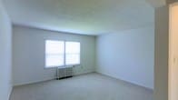 This single-floor space presents a clean, minimalist interior highlighted by a generously sized room visible in the image. The room features neutral-toned wall-to-wall carpeting that contributes to a quiet, comfortable atmosphere underfoot. A textured popcorn ceiling adds a classic architectural character to the space overhead.

Natural light enters through a double-pane window fitted with horizontal blinds, offering both brightness and privacy control. The window's placement along the exterior wall ensures the room receives ample daylight throughout the day. Below the window sits a traditional white cast-iron radiator, indicating a hot-water or steam heating system — a reliable and effective heating solution commonly found in well-established buildings.

The walls are finished in a clean, off-white tone, providing a neutral backdrop that would complement a variety of interior design styles. White baseboards run along the perimeter of the room, adding a crisp, finished detail to the flooring transition. The overall layout of the room is open and unobstructed, offering flexible possibilities for furniture arrangement and room function. The space conveys a straightforward, functional design with practical, enduring features throughout.
