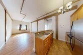 Located at 617 Hiawatha Blvd E in Syracuse, NY, this one-bedroom, one-bathroom unit offers a well-designed single-floor layout with a variety of distinctive architectural details. The open-concept kitchen and living area is a central highlight, featuring warm honey-toned cabinetry, granite-style countertops, a double-basin sink, and a full suite of stainless steel appliances including a refrigerator, range, and microwave. A peninsula counter with bar seating connects the kitchen to the living space, while tile flooring grounds the cooking area. A chandelier overhead adds a touch of character. The living room extends generously from the kitchen, showcasing beautiful hardwood floors, exposed painted brick walls, and structural beam accents that lend the space an industrial-loft aesthetic. Natural light flows in through multiple windows. The spacious bedroom features plush carpeting, tall ceilings, a double-door closet, and windows that invite ample natural light. An additional closet provides extra storage. The bathroom is clean and functional, equipped with a full tub and shower combination. A hallway connects the rooms efficiently, completing this thoughtfully laid-out home with a blend of classic and industrial character.