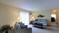 This single-floor space centers around a well-appointed living room designed for comfort and functionality. The room features wall-to-wall carpeting in a neutral tone, contributing to a warm and inviting atmosphere. Large sliding glass doors with vertical blinds serve as a focal point, allowing natural light to fill the space while providing access to an outdoor area. A wall-mounted air conditioning unit above the sliding doors ensures year-round climate control. The living room is furnished with a coordinated grey sectional sofa and matching armchair, accented with navy and white decorative pillows that add visual contrast. A dark wood coffee table with a lower shelf anchors the seating arrangement, complemented by matching end tables and warm-toned floor lamps on either side of the room. Decorative metallic wall art adds a contemporary touch to the otherwise understated wall space. Baseboard heating units are visible along the walls, indicating a practical heating setup. The textured ceiling and light yellow-toned walls create a soft, neutral backdrop that ties the room's elements together cohesively.
