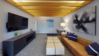 Located at 1 N Main St in Algonquin, Illinois, this single-floor amenity space offers a well-designed, multi-functional environment ideal for socializing and relaxation. The layout flows seamlessly across several distinct areas, each with its own character and purpose.

The lounge area is a standout feature, showcasing a coffered ceiling with warm golden wood beam accents and ambient uplighting that creates an inviting, upscale atmosphere. A wall-mounted flat-screen TV, a leather sofa with coordinating accent pillows, a modular wooden coffee table, and a linear fireplace contribute to a comfortable yet contemporary aesthetic. Neutral gray carpeting runs throughout, tying the spaces together cohesively.

Adjacent to the lounge, a bright and open game and gathering area is bathed in natural light from large, multi-pane windows. A round pedestal table surrounded by sleek mid-century-style chairs provides a casual setting for games, as evidenced by the chess set on display. Teal accent walls add a bold pop of color, while white-framed French doors with glass panes open into an additional room labeled "The Arena," suggesting a dedicated activity or entertainment space.

Throughout the space, curated abstract artwork adorns the walls, reinforcing a modern, polished design sensibility. Ample windows bring in views of the surrounding neighborhood, creating a bright and welcoming environment throughout the day.