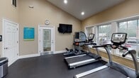 This fitness center is a comprehensively outfitted facility designed to support a wide range of workout routines. The space is laid out across a single floor with a dark rubber flooring surface that provides both durability and safety underfoot. Vaulted ceilings with recessed lighting lend an open, airy feel throughout.

The cardio zone is anchored by multiple True treadmills positioned in front of large windows, allowing natural light to fill the space and offering pleasant outdoor views during workouts. A wall-mounted flat-screen television is conveniently placed for entertainment while exercising.

The strength training area features a multi-function cable machine, a weight bench, and a set of barbells with plates, all set against a striking teal accent wall that adds a bold visual contrast to the warm beige tones found elsewhere in the room. A full rack of dumbbells lines the wall, catering to a variety of strength training needs.

A dedicated functional training station includes a tiered rack stocked with medicine balls, stability balls in multiple sizes, yoga mats, and ab rollers, providing ample tools for core and functional fitness routines. A wall-mounted mirror expands the sense of space and aids in form correction.

Additional amenities include a water fountain and informational signage near the entrance, making this fitness center a practical and inviting environment for daily exercise.
