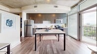 Located on the 18th floor at 848 Spring St NW in Atlanta's vibrant Midtown neighborhood, this one-bedroom, one-bathroom residence offers a thoughtfully designed layout with modern industrial character. The open-concept living and kitchen area is anchored by rich hardwood-style flooring, exposed concrete ceilings, and floor-to-ceiling windows that flood the space with natural light and frame sweeping city views. The kitchen is well-appointed with dark espresso cabinetry, stainless steel appliances including a refrigerator, microwave, and range, and a large central table that doubles as a dining and prep area. The spacious bedroom features plush carpeting, ample room for a full sleeping and workspace arrangement, and a ceiling fan beneath the distinctive exposed concrete ceiling. A dedicated closet provides convenient storage. The bathroom is clean and contemporary, featuring a walk-in glass-enclosed shower with white tile surround and mosaic tile flooring. A hallway connects the main living areas, and an in-unit washer is also present. The overall ambiance blends urban industrial aesthetics with warm, residential comfort, making this a compelling Midtown Atlanta address.