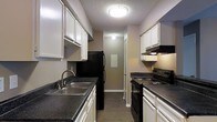 This single-floor space showcases a well-appointed galley-style kitchen as its centerpiece. The kitchen is designed for efficiency, with counters running along both sides of a narrow, elongated corridor layout that maximizes workspace. White cabinetry lines both walls from floor to ceiling, providing generous storage, while dark granite-style countertops offer a striking contrast and durable work surfaces. Black appliances, including a refrigerator, electric range with overhead ventilation, and a double-basin stainless steel sink, lend a cohesive, modern aesthetic to the space. The flooring features wood-style planks that complement the overall color palette. Overhead, a flush-mount ceiling light provides bright, even illumination throughout the kitchen, while an under-cabinet light above the range adds task lighting for cooking. The textured ceiling adds a classic touch to the interior. At the far end of the kitchen, a hallway leads further into the space, hinting at additional rooms beyond. The overall ambiance is clean, practical, and functional, making this kitchen a highly organized and efficient environment suited for everyday living.