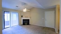 This single-floor space offers a welcoming and functional layout designed for comfortable everyday living. The living area is the centerpiece of the home, immediately drawing attention with its vaulted ceiling that adds a sense of openness and volume to the room. A ceiling fan is centrally positioned, ensuring year-round comfort and airflow throughout the space. Soft, neutral-toned carpeting covers the floor, contributing to a warm and inviting ambiance. A classic fireplace with a sleek black surround and a white mantle serves as a focal point, providing both aesthetic appeal and a cozy gathering spot. Adjacent to the fireplace, a sliding glass door fitted with vertical blinds allows abundant natural light to flood the interior while offering convenient access to the outdoors. The neutral wall tones throughout complement the overall light and airy feel of the space. A secondary door provides additional access or leads to a utility or storage area. The thoughtful combination of architectural details, including the vaulted ceiling, fireplace, and generous natural light, creates a space that feels both spacious and comfortable.