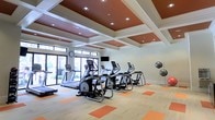 This fitness center offers a thoughtfully designed workout environment that balances both aesthetics and functionality. The space is defined by its striking coffered ceiling, finished in a warm terracotta hue with recessed lighting that provides bright, even illumination throughout. The ceiling's geometric grid pattern adds an architectural elegance rarely found in typical fitness facilities. Underfoot, the flooring features a light wood-toned surface accented with bold orange tile insets, echoing the warm color palette of the ceiling above and creating a cohesive, energizing atmosphere. Large floor-to-ceiling windows and glass doors line one wall, flooding the space with natural light and offering open views to the outdoors, enhancing the sense of openness. The fitness center is outfitted with a full row of modern cardio equipment, including treadmills, elliptical trainers, and a stair climber, all arranged to face the windows. A wall-mounted television provides entertainment during workouts, while medicine ball storage and a wall clock complete the practical amenities. The overall ambiance is clean, bright, and motivating — a well-appointed facility suited for a focused and enjoyable fitness experience.