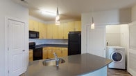 The Waldron is a thoughtfully designed 2-bedroom, 2-bathroom residence laid out on a single floor. The open-concept living and dining area features rich, dark wood-style flooring that flows seamlessly toward the kitchen, creating a cohesive and welcoming atmosphere. The kitchen is outfitted with light maple cabinetry, dark countertops, black appliances including a refrigerator, range, and built-in microwave, and a curved island with an undermount sink — illuminated by elegant pendant lighting above. Both bathrooms share a consistent design aesthetic, featuring warm wood-tone vanity cabinetry, dark countertops, undermount sinks with modern fixtures, large mirrors, and tiled tub-shower combinations set against wood-style flooring. The two bedrooms are carpeted for comfort and offer generous proportions, with windows that bring in natural light. One bedroom includes a walk-in closet equipped with wire shelving, providing ample storage. Additional storage is available throughout the unit, including a louvered-door closet near the kitchen. The overall design balances contemporary finishes with functional, everyday living in a clean, neutral palette.