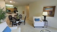 This single-floor residence offers an open-concept design that seamlessly integrates the living, dining, and kitchen areas into one cohesive space. The living area features plush wall-to-wall carpeting in a neutral tone, creating a warm and comfortable atmosphere. A white upholstered armchair accented with a blue decorative pillow anchors the seating area, complemented by a side table with a classic lamp. The dining area accommodates a glass-top table surrounded by black wooden chairs with cushioned seats, providing an elegant yet functional spot for meals. Adjacent to the dining space, the kitchen is visible with warm wood cabinetry and overhead task lighting, offering practical cooking amenities. The walls throughout are finished in a soft, neutral palette that enhances the sense of openness and brightness in the space. A closeted door and hallway visible in the background suggest additional storage and private rooms beyond the main living area. The overall ambiance is clean, understated, and well-organized, with a consistent design language that balances comfort and functionality throughout the home.