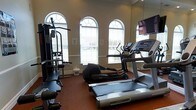 Located at 100 Saddle Cove Road in Bohemia, NY, this fitness center offers a well-appointed single-floor space designed to support a comprehensive workout experience. The room is characterized by its classic architectural details, including tall arched windows with decorative transoms that flood the interior with natural light, creating an open and inviting atmosphere. Crown molding and clean wall finishes contribute to an upscale aesthetic that sets this facility apart from a typical gym environment. The space is equipped with a variety of exercise machines, including a treadmill, elliptical trainer, and a multi-station strength training machine, catering to both cardio and resistance training needs. A wall-mounted television is positioned for convenient viewing during workouts. Large mirrors along the walls enhance the sense of space while allowing users to monitor their form. The rubber flooring provides both safety and durability underfoot. The combination of elegant design elements and functional fitness equipment makes this a practical yet refined space suited for residents or members seeking a convenient and comfortable place to maintain an active lifestyle.