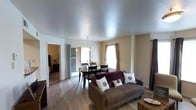 This single-floor residence offers a well-organized, open-concept layout that seamlessly connects the living and dining areas, creating a comfortable and functional living environment. The living area is anchored by a cozy sofa accented with decorative throw pillows and is finished with warm hardwood-style flooring that extends throughout the main space. A textured ceiling adds a classic touch to the interior.

The dining area sits adjacent to the living space and is defined by a dark-toned dining table and chairs, illuminated by a pendant light fixture hanging above. Sliding glass doors lead directly from the dining area to an outdoor space, flooding the interior with natural light and providing convenient outdoor access. A window with curtain panels in the dining and living areas further enhances the bright, airy atmosphere.

An open doorway from the main living area provides a view into what appears to be a bedroom, contributing to the flowing connectivity of the floor plan. The neutral, warm-toned walls throughout complement the light flooring and create a clean, inviting ambiance. Overall, this space is designed for comfortable everyday living, with practical features and a cohesive aesthetic that ties each area together naturally.