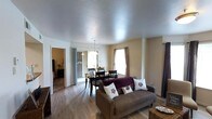 This single-floor residence offers a well-organized, open-concept layout that seamlessly connects the living and dining areas, creating a comfortable and functional living environment. The living area is anchored by a cozy sofa accented with decorative throw pillows and is finished with warm hardwood-style flooring that extends throughout the main space. A textured ceiling adds a classic touch to the interior.

The dining area sits adjacent to the living space and is defined by a dark-toned dining table and chairs, illuminated by a pendant light fixture hanging above. Sliding glass doors lead directly from the dining area to an outdoor space, flooding the interior with natural light and providing convenient outdoor access. A window with curtain panels in the dining and living areas further enhances the bright, airy atmosphere.

An open doorway from the main living area provides a view into what appears to be a bedroom, contributing to the flowing connectivity of the floor plan. The neutral, warm-toned walls throughout complement the light flooring and create a clean, inviting ambiance. Overall, this space is designed for comfortable everyday living, with practical features and a cohesive aesthetic that ties each area together naturally.