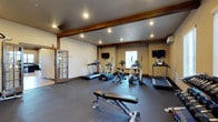 This fitness center offers a comprehensive workout environment designed to support a wide range of exercise routines. The space is defined by its clean, functional layout and distinctive architectural character, including exposed wooden beam accents that add a warm, rustic touch to the otherwise modern interior. The dark rubberized flooring provides a durable, slip-resistant surface throughout the facility.

The cardio area is well-appointed with multiple treadmills, elliptical trainers, and a stair climber, all positioned for convenient use. Two wall-mounted flat-screen televisions are strategically placed to keep users entertained during workouts. A wall-mounted split air conditioning unit ensures a comfortable climate year-round.

A dedicated free weights section includes a full dumbbell rack with an extensive range of weights, medicine balls, a stability ball, and an adjustable weight bench, catering to both strength training and functional fitness. Natural light filters in through windows fitted with horizontal blinds, creating a bright and welcoming atmosphere.

Storage shelving and organizational cubbies provide practical space for personal items and additional equipment. The room is accessed through a glass-paneled French door, adding a refined aesthetic detail. Recessed ceiling lighting throughout ensures the space is well-illuminated at all times, making this fitness center a fully equipped and inviting facility for daily exercise.