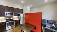 This 2-bedroom, 2-bathroom unit is thoughtfully arranged on a single floor, offering a functional and well-connected layout. The kitchen is a standout feature, equipped with dark espresso cabinetry, stainless steel appliances including a refrigerator and range, and a stone-tile backsplash that adds texture and visual interest. Light countertops provide a clean contrast to the rich cabinetry tones, while recessed lighting ensures the space is well-illuminated throughout.

A bold red partition wall serves as a defining architectural element, visually separating the kitchen and dining area from the living space beyond. This creative room divider adds a strong pop of color and gives the open-plan layout a sense of defined zones without closing off the space. The dining area accommodates a dark wood table, complementing the kitchen's color palette.

The living area, visible beyond the partition, features a comfortable seating arrangement and continues the warm, cohesive aesthetic. Hardwood flooring runs throughout, adding warmth and continuity. The overall ambiance is modern and practical, with design choices that maximize the use of the available space.