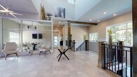 This two-story community building masterfully blends modern design with warm, rustic elements throughout its generously scaled common areas. The dramatic main hall immediately impresses with soaring double-height ceilings, large-format tile flooring, and bold dark wood structural columns that anchor the space with character. A sweeping staircase with black metal railings connects both floors, complemented by a stainless steel elevator for convenient accessibility. Upper-level clerestory windows draw in abundant natural light, enhancing the open, airy atmosphere.

The main level also features a glassed-in lounge area, creating an intimate retreat within the expansive common space. A comfortable sitting lounge on the lower level offers plush seating arrangements, textured accent walls, and stylish décor, ideal for relaxed gatherings. A dedicated dining room provides round tables with garden views through large windows, while the communal kitchen is outfitted with extended countertops, gray shaker cabinetry, stone-look surfaces, wood-look flooring, and pendant lighting. A hallway corridor features a striking blue tile accent wall alongside a water station and restroom access, adding a refined detail to the circulation path. Throughout, warm wood flooring, recessed lighting, and cohesive neutral tones create a welcoming and functional environment.