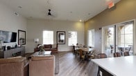 The clubhouse at 3875 Main in Chula Vista is a thoughtfully designed single-floor community gathering space that seamlessly blends comfort and functionality. The open-concept layout connects the main lounge, dining area, and kitchen, creating a welcoming environment ideal for both casual gatherings and organized events.

The lounge area features soaring ceilings, recessed lighting, and a ceiling fan, contributing to an airy and comfortable atmosphere. Rich brown leather seating is arranged around a wood-and-metal coffee table, complemented by a wall-mounted flat-screen TV and a media console. Framed artwork adds a warm, curated touch to the neutral-toned walls. Large windows with slatted blinds flood the space with natural light throughout the day.

The kitchen is well-appointed with dark espresso cabinetry extending to the ceiling, providing ample storage. A decorative patterned tile backsplash adds visual interest, while light quartz countertops and a central island offer generous prep and serving space. Stainless steel appliances, including a French door refrigerator and dishwasher, complete the fully functional setup.

The dining area accommodates multiple tables with metallic bistro-style chairs, suited for community dining or meetings. Glass-paned French doors connect the interior to an outdoor patio area, further enhancing the sense of openness. Warm wood-look flooring runs throughout the entire space, tying together the modern yet inviting aesthetic.