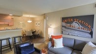This single-floor residence offers an open-concept layout that seamlessly connects the living and dining areas, creating a cohesive and functional flow throughout the space. The living area is furnished with a plush gray sofa accented by vibrant orange and white throw pillows, complemented by a sleek leather chair. A table lamp with a chrome base provides warm ambient lighting, while a bold urban-themed artwork depicting a classic transit station serves as a striking focal point on the neutral-toned wall. The dining area, partially separated by a structural column, features a round dark wood table surrounded by upholstered chairs in a complementary neutral tone. A pendant light fixture hangs above the dining table, casting a warm, inviting glow over the space. Framed artwork adorns the dining area walls, adding a refined touch. The flooring transitions between dark hardwood in the dining area and a patterned area rug in the living space, adding texture and visual interest. The overall ambiance is warm and contemporary, blending comfort with a modern urban aesthetic throughout.
