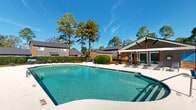 Located in Aiken, South Carolina, this outdoor pool area offers a well-appointed recreational retreat designed for relaxation and leisure. The centerpiece is a generously sized swimming pool with clear, blue-green water, featuring a shallow end clearly marked for safety. The pool's clean geometric shape is complemented by a wide, light-colored concrete sundeck that wraps around the perimeter, providing ample space for sunbathing and socializing.

Numerous lounge chairs are arranged throughout the deck, offering plentiful seating for guests to unwind in the South Carolina sunshine. A covered pavilion structure with a pitched roof and exposed wood beam detailing anchors one side of the area, providing shaded seating and serving as a functional gathering space. The pavilion's design incorporates brick and modern gray siding, blending classic and contemporary elements.

Round patio tables with chairs are also distributed across the deck, ideal for outdoor dining or casual conversation. The entire pool area is enclosed by a fence and bordered by manicured hedgerows and lush, mature trees that provide natural privacy and shade. The surrounding canopy of tall pines and deciduous trees enhances the serene, park-like atmosphere, making this an inviting outdoor amenity in the heart of Aiken.
