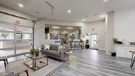 This well-appointed amenity facility spans a single floor and is thoughtfully divided into two distinct zones: a welcoming lounge area and a fully equipped fitness center. The lounge area makes a strong first impression with its wide-plank wood-look flooring in cool grey tones, high ceilings, and an abundance of natural light streaming through large windows dressed with floor-length drapes. The space is furnished with modern seating arrangements, including plush sofas and accent chairs, accented by glass-and-gold coffee tables and lush indoor plants. A dedicated coffee bar area features white cabinetry, a sink, and a dining table with sleek black chairs — ideal for casual gatherings or focused work sessions. A large pass-through window and a retractable garage-style door connect the lounge seamlessly to the fitness center, creating an open, airy atmosphere. The fitness center is outfitted with a comprehensive array of cardio equipment, including treadmills, stationary bikes, and ellipticals, alongside weight machines and free weights. Floor-to-ceiling mirrors visually expand the space, while a bold motivational typographic wall mural adds energy and character. Recessed and panel lighting throughout ensures a bright, functional environment in both areas.
