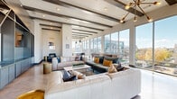 Located in the heart of Nashville's Music Row neighborhood, this sophisticated space delivers an impressive blend of modern design and urban energy. The expansive living room is anchored by soaring ceilings with distinctive exposed dark wood beams and wide-plank hardwood floors that run throughout. A dramatic wall of floor-to-ceiling windows floods the interior with natural light and frames sweeping views of the Nashville skyline, creating an airy, open atmosphere. The living area is generously proportioned, comfortably accommodating multiple seating arrangements alongside a full-size shuffleboard table, making it equally suited for relaxed lounging or lively social gatherings. A built-in media and display wall with custom slate-blue cabinetry adds a music-inspired accent, nodding to the space's iconic Music Row address. A wet bar area with pendant lighting and bar seating provides a functional entertaining hub. The space flows seamlessly onto a sprawling balcony finished with wood-tile decking and glass-panel railings, offering open-air seating with unobstructed panoramic views of the Nashville cityscape and beyond. A covered outdoor area with ceiling fans extends the entertaining space further, making this an exceptional venue for experiencing the best of Nashville from above.