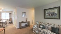 This single-floor space presents a welcoming living area characterized by its clean, neutral aesthetic. The room features wall-to-wall carpeting in a warm beige tone, contributing to a comfortable and inviting atmosphere. Soft, light grey walls provide a versatile backdrop throughout the space, complemented by standard-height ceilings that maintain an intimate feel.

The living area is thoughtfully arranged, with a floral-patterned armchair paired with decorative candle-style floor lamps, adding a touch of classic charm. A framed artwork piece adorns one wall, enhancing the room's personality. A rustic wooden console table, styled with a small mirror, decorative vases, and dried botanicals, serves as a practical and attractive focal point near the entryway transition.

A floor-level ventilation register is visible along the wall, indicating a central HVAC system. The layout suggests an open flow between the living area and adjacent spaces, visible through a doorway leading further into the home. The overall ambiance is understated and functional, with a traditional design sensibility throughout.