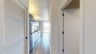 Located in Chicago's vibrant Division Street corridor, this one-bedroom, one-bathroom unit showcases a clean, contemporary design with industrial-modern touches throughout. The open-concept living and kitchen area is defined by striking exposed concrete ceilings and light wide-plank hardwood flooring, creating a refined urban aesthetic. The galley-style kitchen is outfitted with stainless steel appliances, a gas range with oven, a dark tile backsplash, and sleek gray cabinetry with under-cabinet lighting, offering both style and functionality. Floor-to-ceiling windows flood the living space with abundant natural light and frame sweeping elevated views of the Chicago skyline and surrounding neighborhoods. An exposed concrete structural column adds to the industrial character of the space. The bedroom continues the minimalist theme with the same hardwood flooring, a large floor-to-ceiling window with city views, and a built-in closet. A hallway connects the main living areas to the well-appointed bathroom, which features large-format tile flooring, a dark wood vanity with a white countertop, a wide mirror, and a full soaking tub with a dark tile surround and handheld shower. The overall ambiance is modern, light-filled, and thoughtfully designed for urban living.