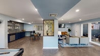 Located at 7510 Brompton St in Houston, TX, this thoughtfully designed single-floor space offers a cohesive and visually striking interior defined by a cool blue-and-gold color palette. The open layout seamlessly connects multiple functional zones, creating a fluid and inviting environment throughout.

The lounge area serves as a comfortable gathering space, featuring a plush navy blue sofa, a patterned area rug in blue and grey tones, a wall-mounted flat-screen TV, and a warm wood credenza. Tall decorative plants add a touch of greenery against the soft blue walls. A standout gold geometric lattice partition divides the lounge from adjacent areas, functioning as both a design focal point and an elegant room separator.

The dining zone offers upholstered navy banquette seating alongside glass-top tables and modern chairs, illuminated by dual gold pendant lights. A horizontal window brings in natural light and views of the surrounding outdoors.

The wet bar area is finished with deep navy cabinetry, a mosaic tile backsplash, open shelving, and a beverage cooler — ideal for entertaining. Adjacent to this, a game room features a full-size pool table, rounding out the entertainment amenities.

Herringbone-patterned hardwood flooring flows consistently across the entire space, tying together each zone with a refined, modern aesthetic. Recessed lighting throughout ensures a well-lit, polished atmosphere.