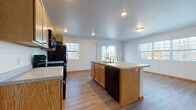 This single-floor unit is designed with an open-concept layout that seamlessly connects the kitchen and living area, creating a spacious and functional environment. The kitchen is well-equipped with light oak cabinetry, black appliances including a refrigerator, electric range, and built-in microwave, alongside light-toned countertops. A central island with an integrated sink and dishwasher serves as a practical workspace and natural divider between the kitchen and the open living space. Multiple windows throughout allow natural light to fill the common areas, offering views of the surrounding exterior.

Wood-look flooring runs consistently throughout the unit, providing a warm, cohesive aesthetic. The bedrooms are comfortably sized, each featuring closet storage and ceiling fans or overhead lighting. One bedroom includes a dedicated closet with built-in shelving. The unit includes two bathrooms: one with a full tub-shower combination and another with a standalone walk-in shower stall. Both bathrooms feature wood-look flooring and clean white fixtures. The overall design is practical and neutral, with white walls and warm wood tones creating a functional living space.