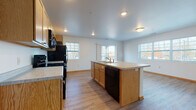 This single-floor unit is designed with an open-concept layout that seamlessly connects the kitchen and living area, creating a spacious and functional environment. The kitchen is well-equipped with light oak cabinetry, black appliances including a refrigerator, electric range, and built-in microwave, alongside light-toned countertops. A central island with an integrated sink and dishwasher serves as a practical workspace and natural divider between the kitchen and the open living space. Multiple windows throughout allow natural light to fill the common areas, offering views of the surrounding exterior.

Wood-look flooring runs consistently throughout the unit, providing a warm, cohesive aesthetic. The bedrooms are comfortably sized, each featuring closet storage and ceiling fans or overhead lighting. One bedroom includes a dedicated closet with built-in shelving. The unit includes two bathrooms: one with a full tub-shower combination and another with a standalone walk-in shower stall. Both bathrooms feature wood-look flooring and clean white fixtures. The overall design is practical and neutral, with white walls and warm wood tones creating a functional living space.