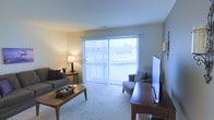This single-floor space offers a comfortable and inviting atmosphere, centered around a well-appointed living area. The room is anchored by large sliding glass doors dressed with vertical blinds, allowing an abundance of natural light to fill the space while providing outdoor views of surrounding greenery. The plush wall-to-wall carpeting adds warmth underfoot and contributes to the cozy ambiance throughout. The living area is generously proportioned, comfortably accommodating a full seating arrangement that includes a sofa and accent chair, paired with a classic wooden coffee table and a coordinating side table topped with a warm-toned lamp. A flat-screen television setup is positioned opposite the seating area, creating a practical and functional entertainment space. The neutral wall tones and soft lighting lend the room a calm, understated aesthetic that feels both livable and welcoming. The layout is open and uncluttered, allowing for easy movement throughout the space. Overall, this single-floor residence combines practical design with a relaxed, comfortable atmosphere, making it a functional and pleasant place to call home.